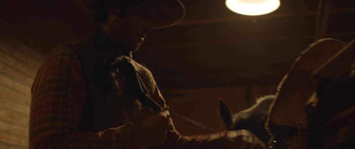 A man wearing a cowboy hat tightens a strap as he saddles a horse in a stable