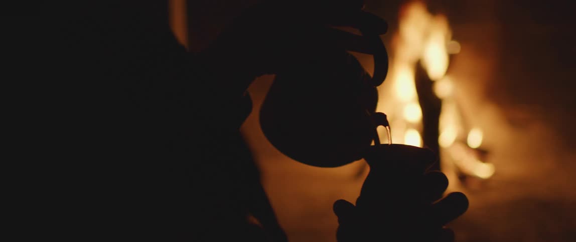 A person pouring hot water from a kettle into a yerba mate gourd in front of a camp fire