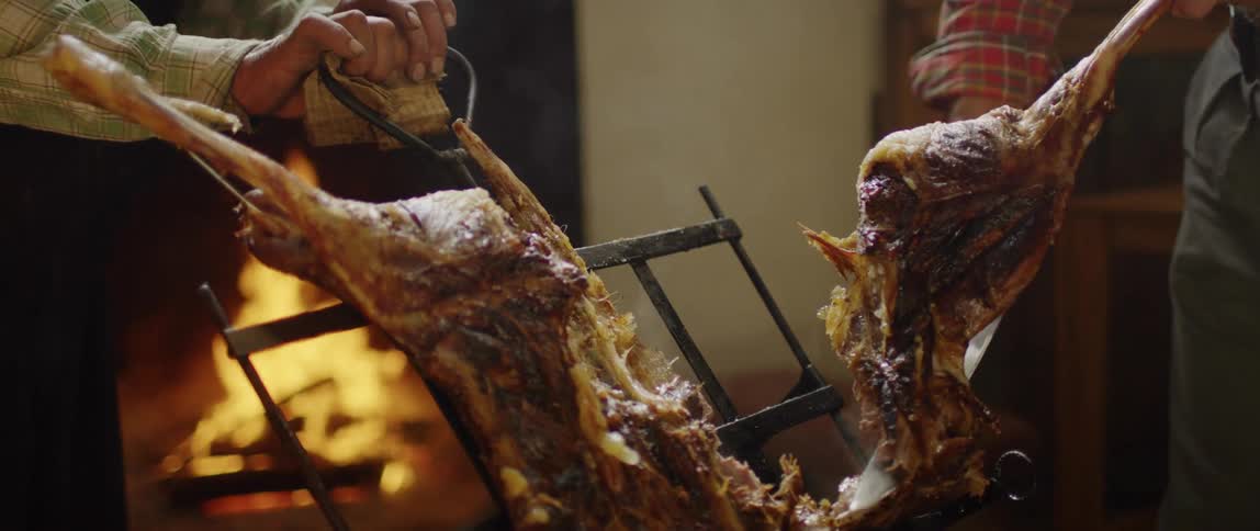 One person holds rack with cooked meat on it while another person cuts off a leg there is a fire in the background
