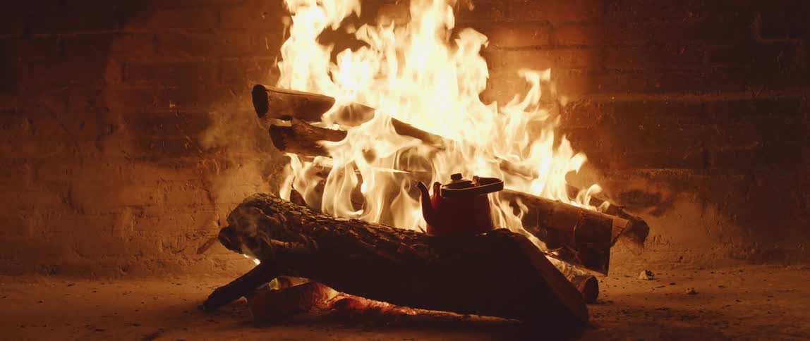 A red kettle sits on a log heating with flames and embers behind it