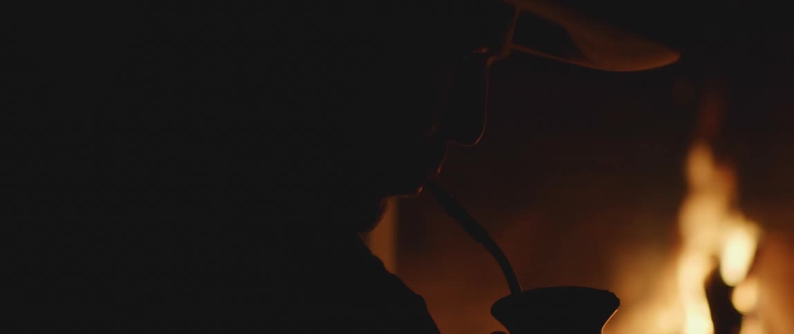A man sits next to a fire and drinks out of a straw