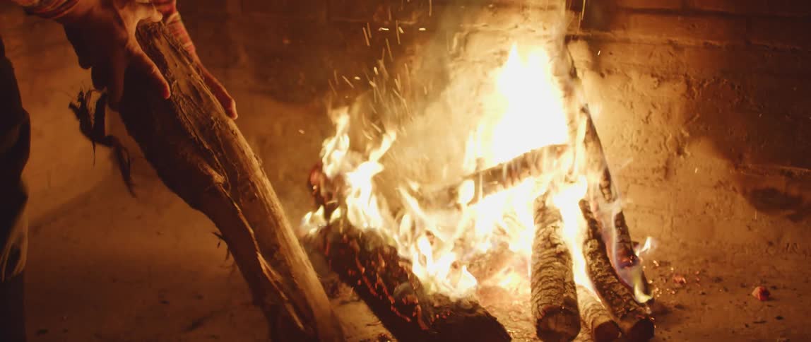 A man places a wood log onto a burning pile of logs in a fireplace