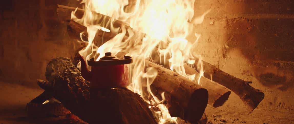 Metal kettle placed over a log in a campfire near a brick wall at night