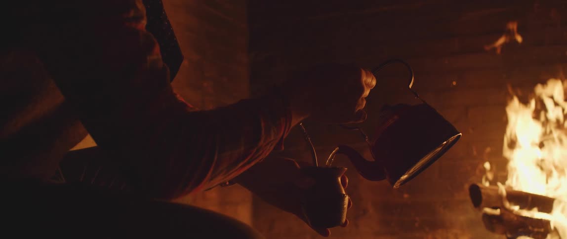 A person pours liquid from a teapot into a cup while sitting in front of a fire