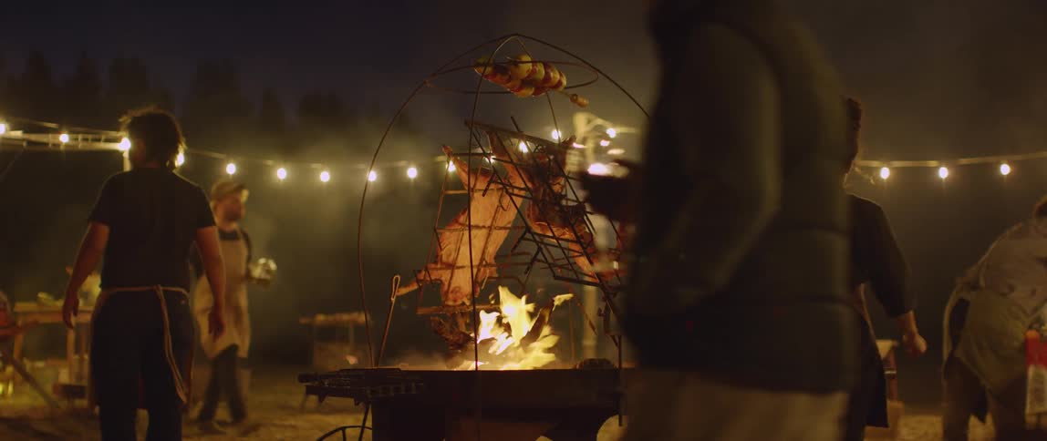 A group of people walking around a camp fire that has roasting meat cooking