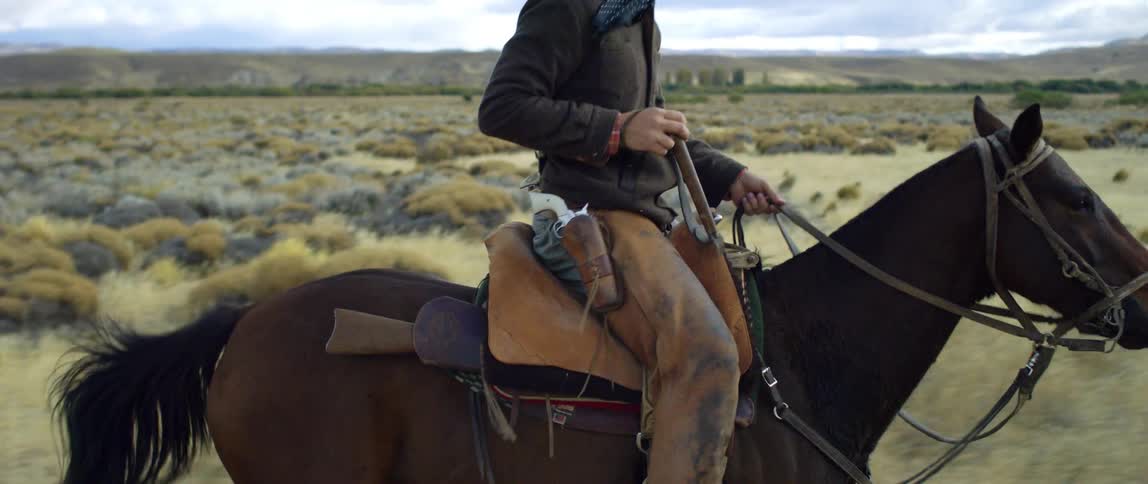 A man rides on horseback on a saddle with a rifle and holding reins during the day