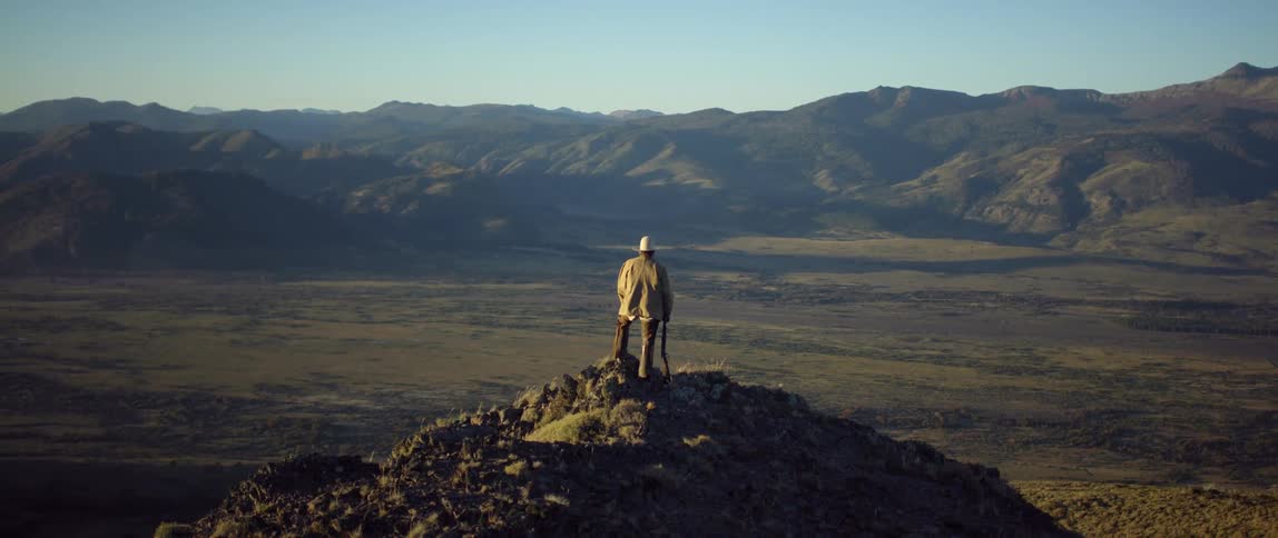 Man wearing cowboy hat and jacket stands on hilltop facing a mountain range