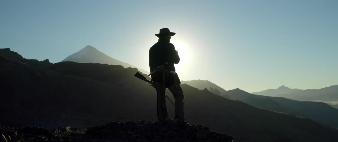 A man wearing a cowboy hat smokes a cigarette while holding a rifle looking over a mountain range