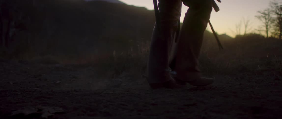 A rancher wearing tall cowboy boots walks through a trail in a field