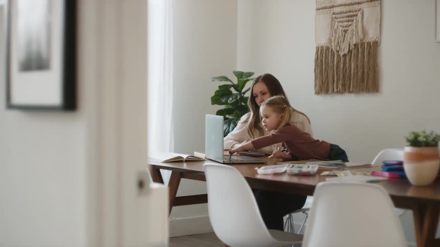 A child sitting at a dining room table next to her mom reaches over to type on a laptop