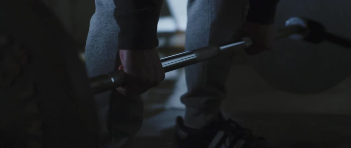 A weightlifter in a sweatsuit lowers a barbell to the floor in a dimly lit room