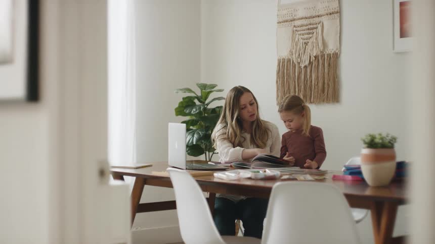 A mother sits at home with her daughter and helps her read a book