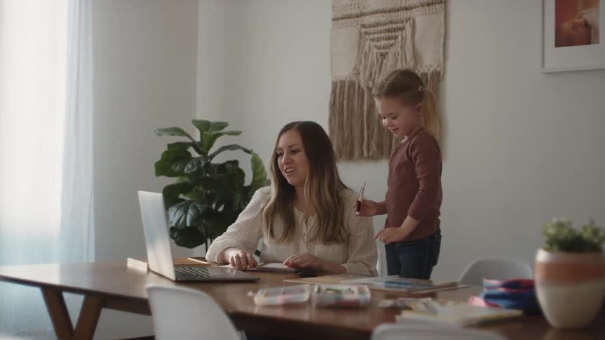 Mother and daughter sit at the table and make a video call on laptop in a room