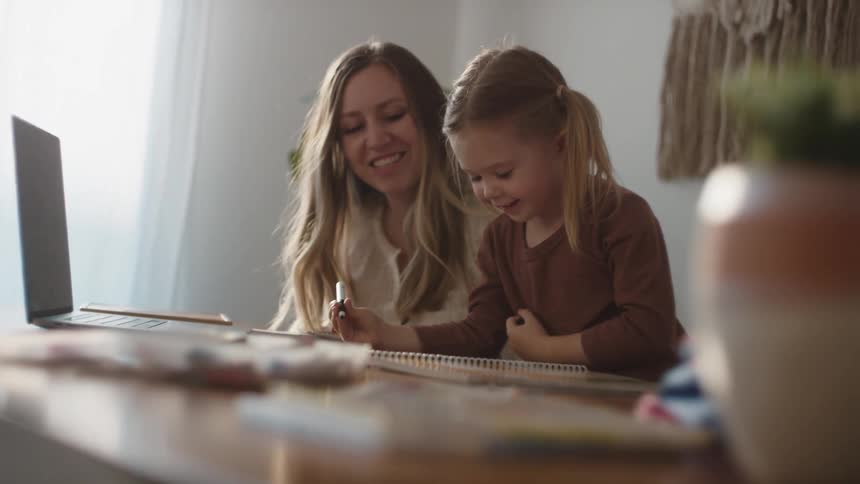 A young girl sits at a table drawing with her mother kneeling beside her