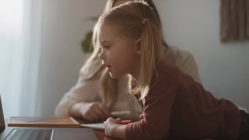 A young toddler girl presses a key on a laptop with her mother kneeling beside her