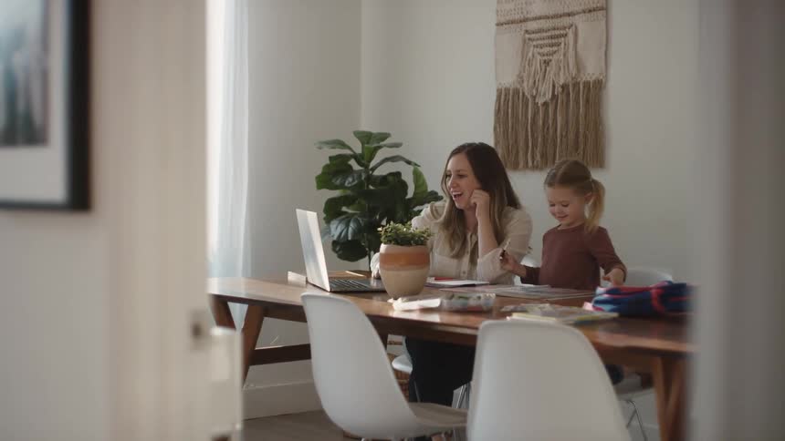 A woman and her daughter wave at the camera of a laptop sitting on a table in a house during the day