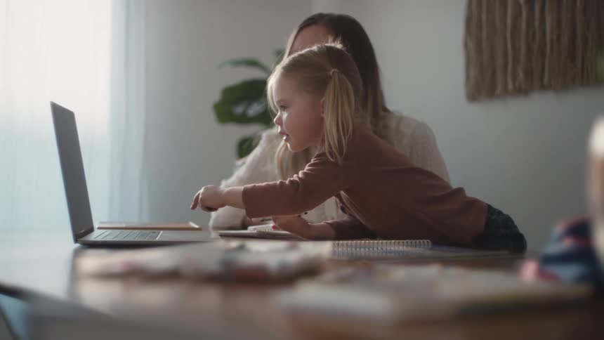 A young girl taps the touchpad on a laptop while her mother watches her from the side