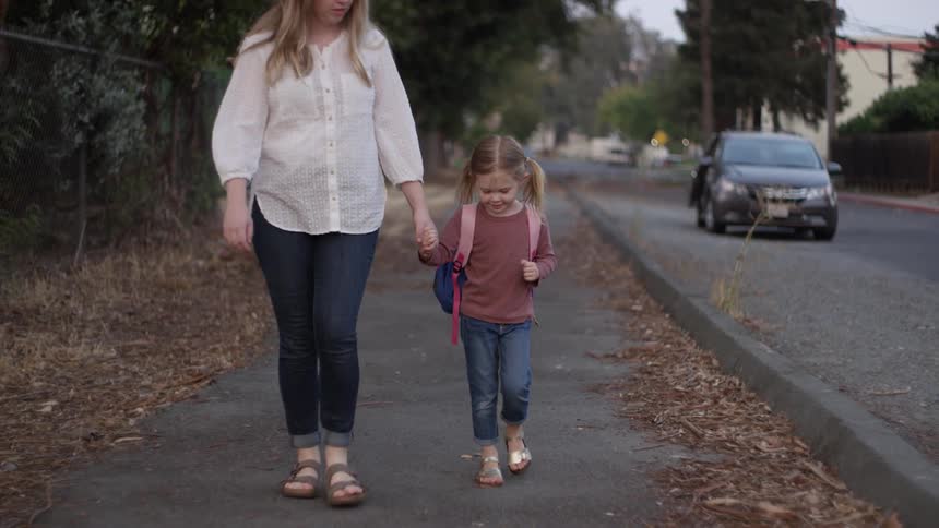 A mother and daughter hold hands walking down a sidewalk with the daughter wearing a back pack