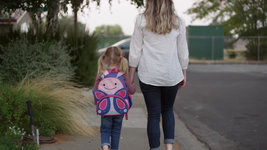 Mother walks her daughter holding hands to school in the sidewalk on a neighborhood