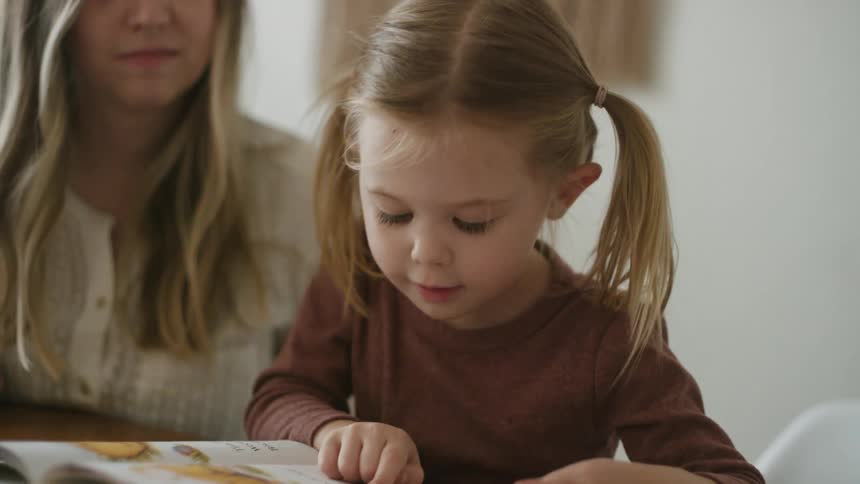 Mother watches as daughter reads book while they sit at table