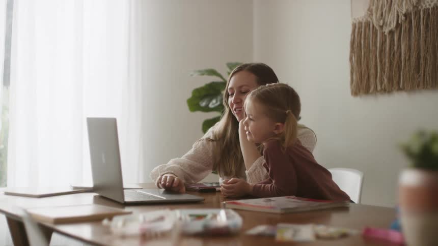Mom and daughter sit at a table as they look at the screen of a laptop during the day