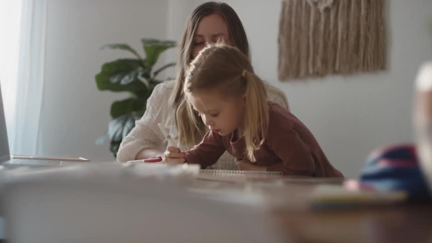 A woman kneels beside her toddler girl who is drawing at a table