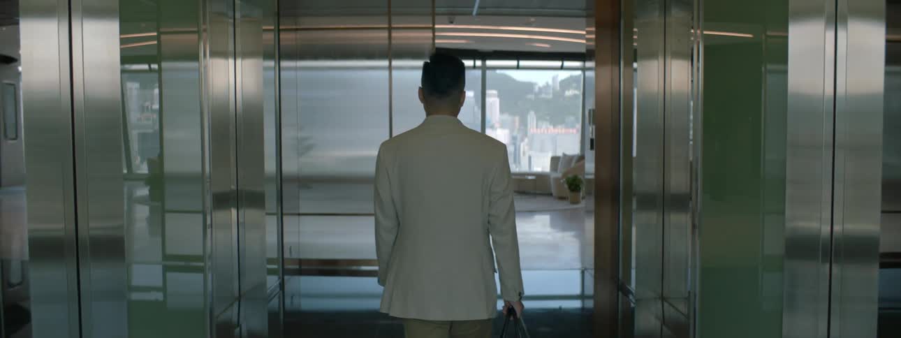 A man wearing a white suit exits an elevator into a seating area where he is greeted by a woman wearing a blue suit