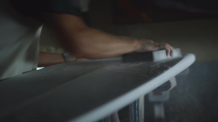 Man sands down surfboard on stand in front of window in dark room