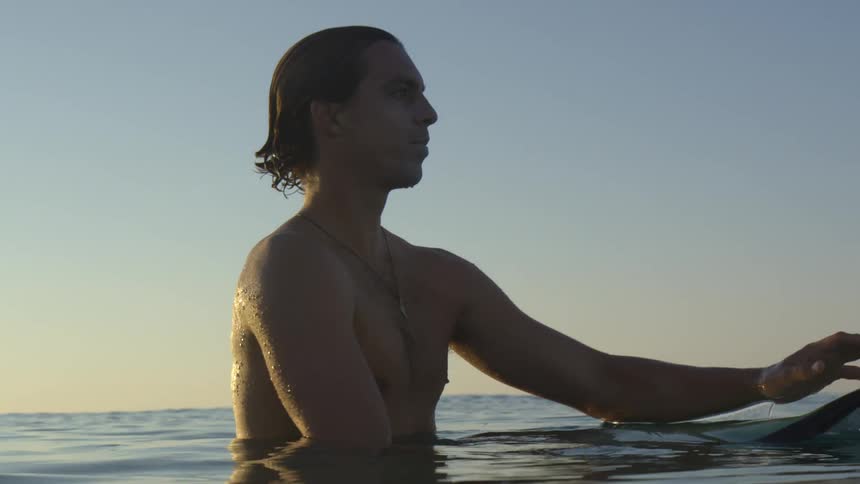 A young man in the ocean floats on his surfboard while taking in his surroundings