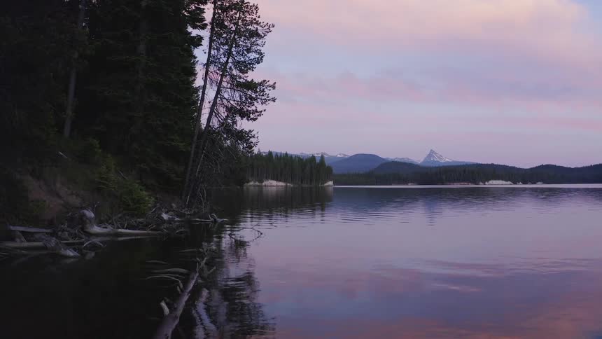 Lake at sunset with trees and dirt hills lined along the shore and mountains in the distance