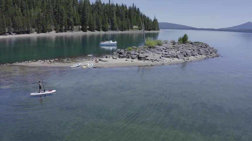 A person paddle boarding on a lake while people and an animal run on an island with trees the sky and clouds in the background