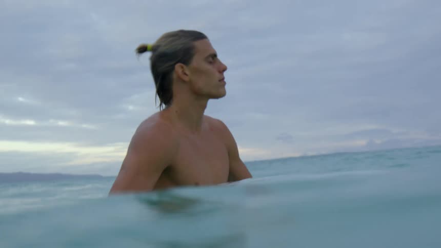 A young man walks through chest deep ocean water under a cloudy daytime sky