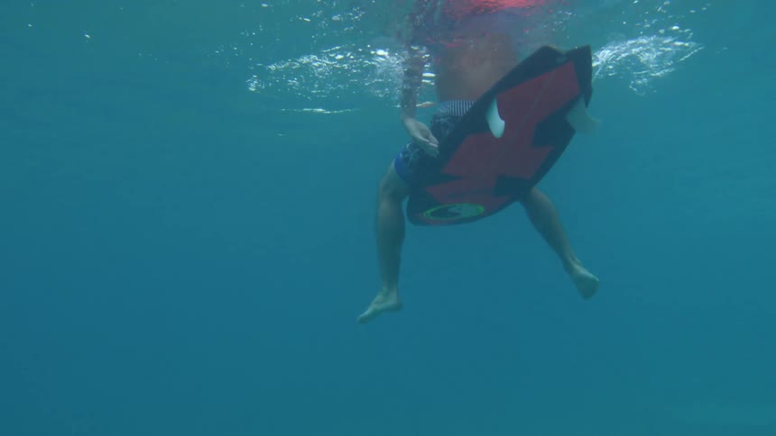 Man with swimming shorts sits on red and black surfboard in the sea water