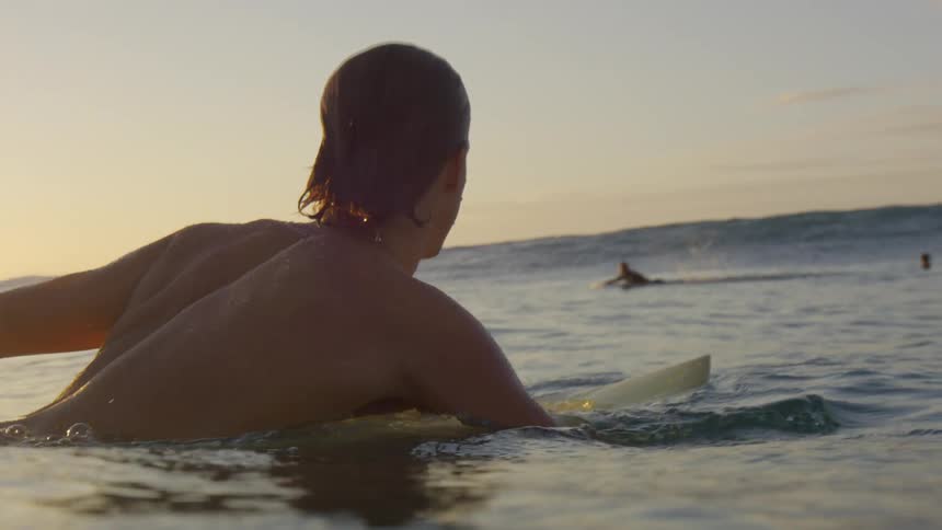Young fit man swimming in the ocean over a surfboard at sunset