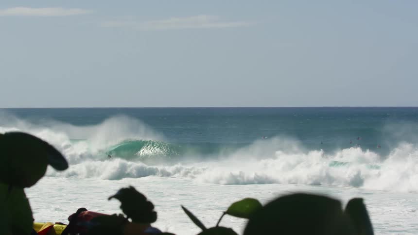 Many surfers are in the water of the ocean while some ride a crashing wave on a sunny day