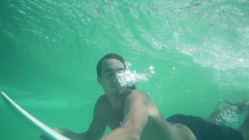 A young man divers underwater with his surfboard and exhales air from his nose as he slowly rises towards the surface
