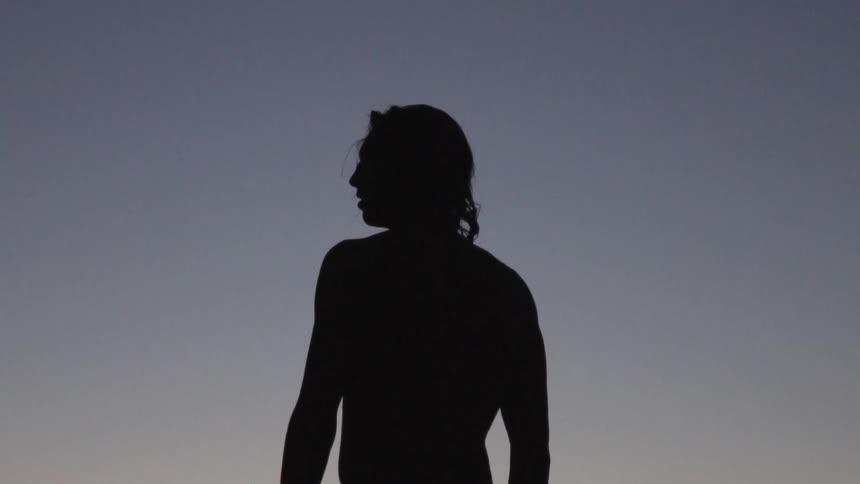 Man with long hair is silhouetted against the sky on the beach at dusk