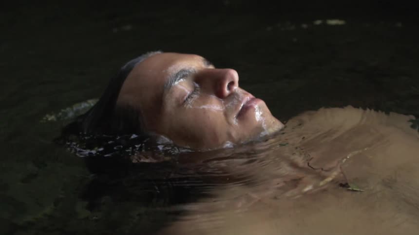A shirtless man lying underwater in a pool of water outdoors slowly sits up so that his head emerges from the water