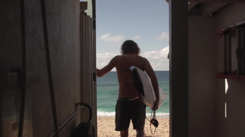 A man carrying a surfboard opens and door and walks out on to the beach