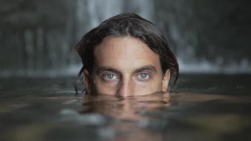 A man with the top half of his head poking out from a pool of water at the base of a waterfall stares into the camera