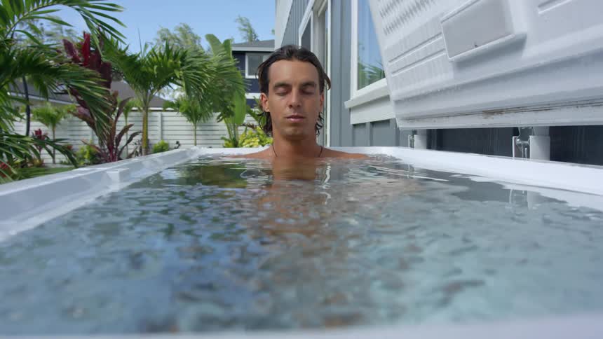 Man sits in tub of ice water in front of buildings palm trees and grass