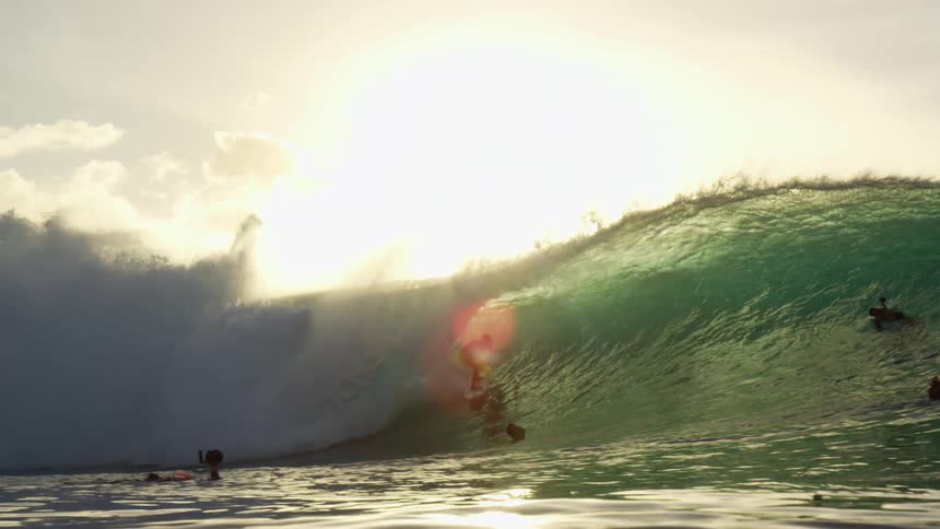 A man surfs under the crashing waters of a wave as swimming camera operators film him from the surrounding sea
