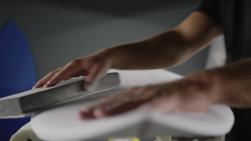 A man sands a surfboard and smooths it with his hands in an indoor setting