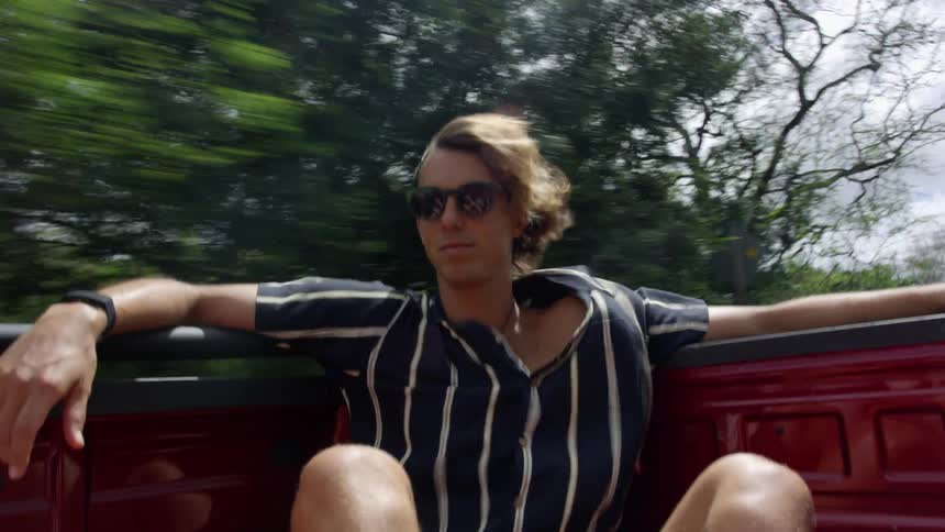 Man sits in bed of red truck driving past woods smiling as wind blows his hair