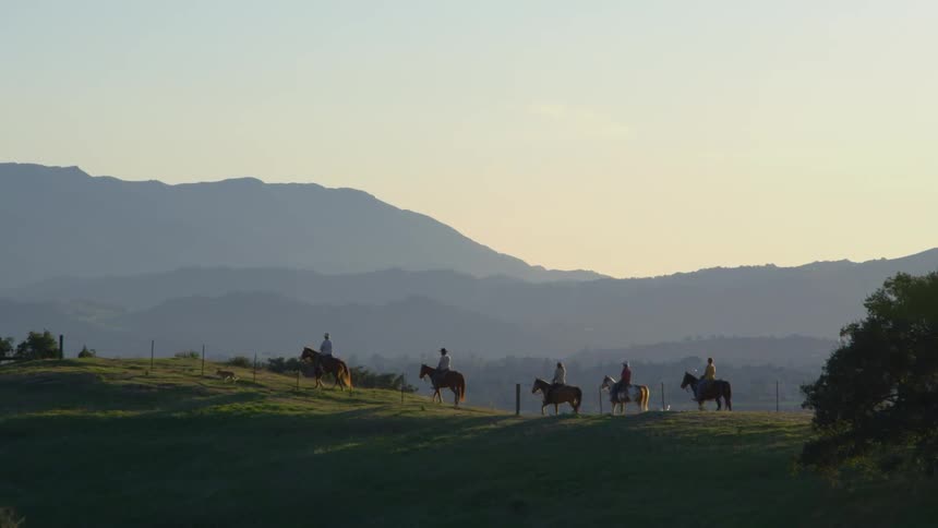 Dogwalks ahead of group of horse riders paralleling fence as sky over foggy mountain is slightly tinted