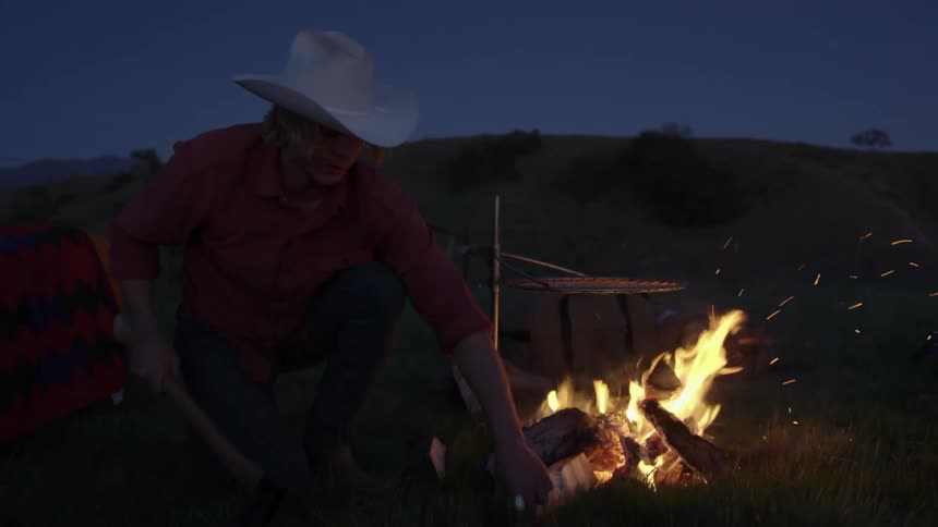 Cowboy chops firewoodnext to campfire near hill
