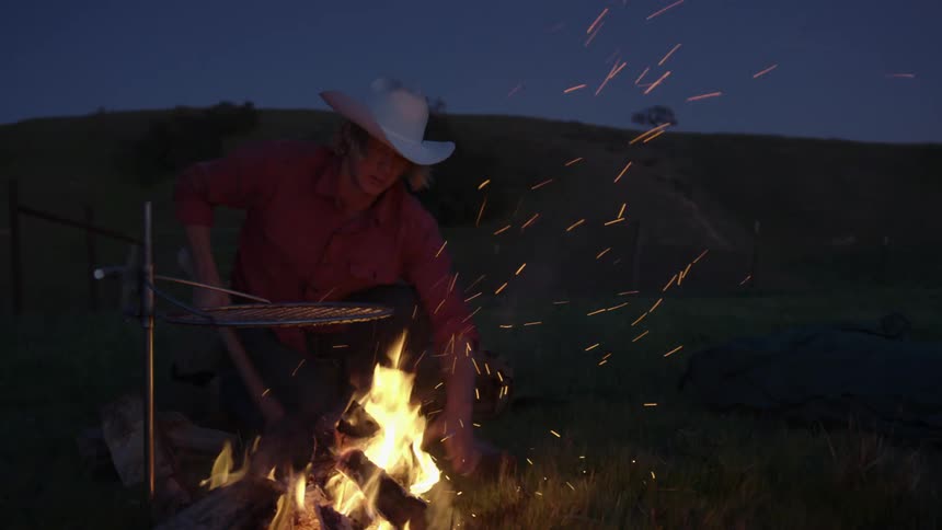 Man with a cowboy hat and a red shirt cuts a wood with an ax next to the campfire at dusk