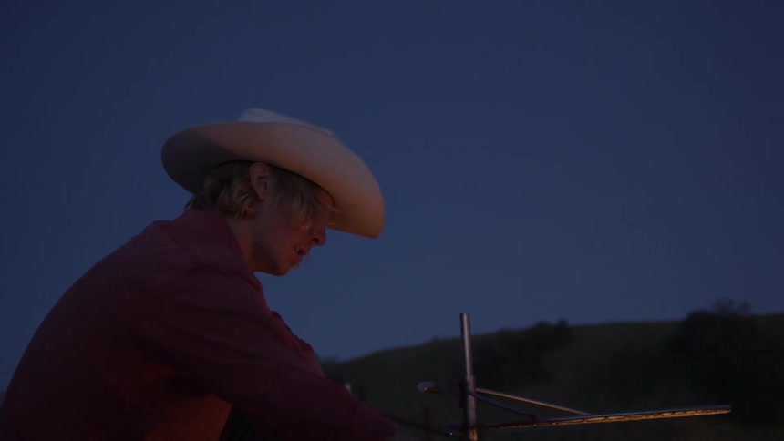 Man wearing a cowboy hat sitting in front of a campfire putting pieces of wood into it at nighttime