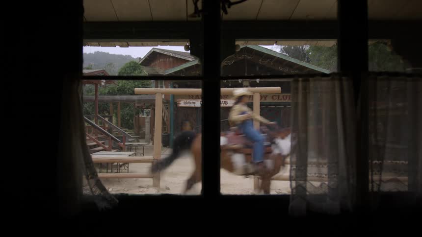 Woman rides by outside of cowboy stage on a brown and white horse