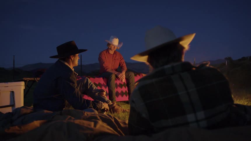 Men wearing cowboy gear sitting around a campfire outdoors and talking amongst each other